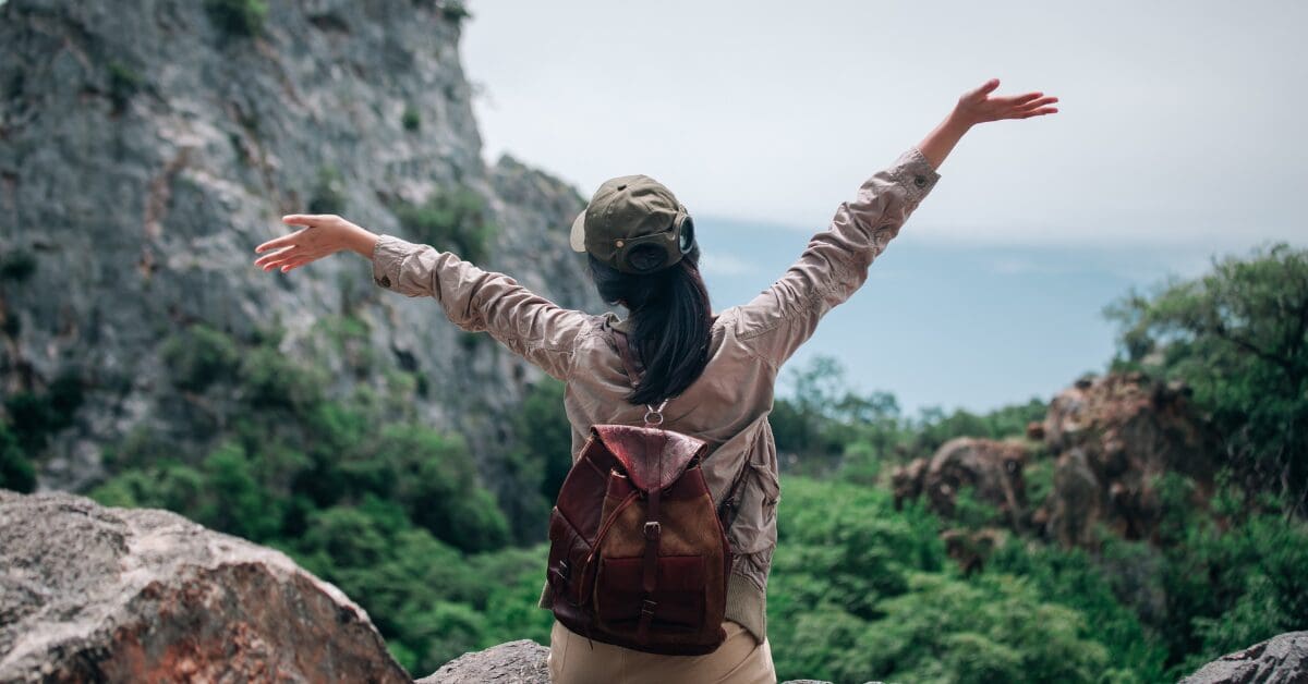 Solo woman on a hiking trip, arms outstretched in freedom and joy, surrounded by mountains and lush greenery—celebrating the power of solo travel.