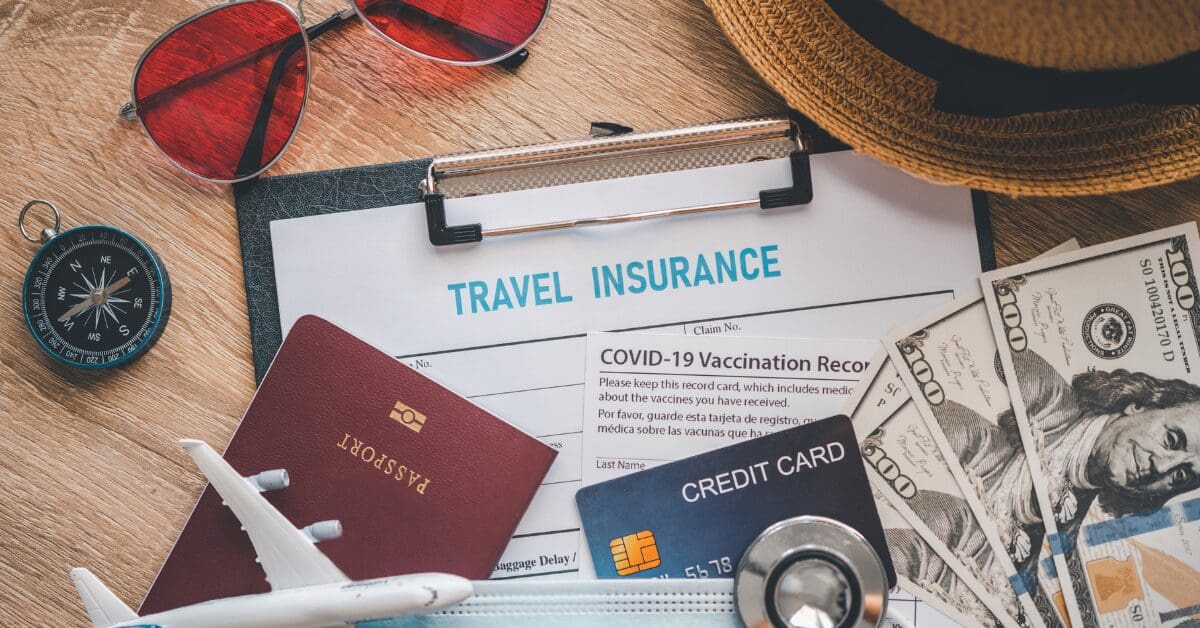Flat lay of travel essentials including a travel insurance form, passport, credit card, cash, vaccination card, sunglasses, and compass—emphasizing smart trip preparation.