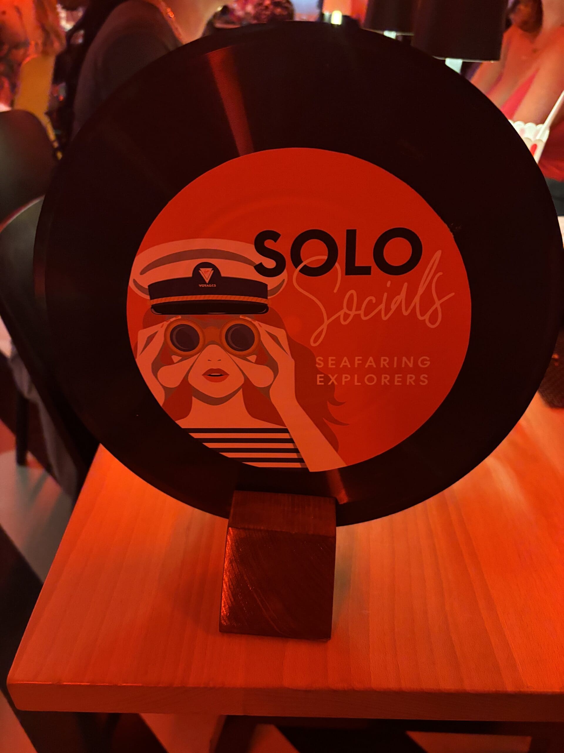 Decorative vinyl record-style sign reading “Solo Socials: Seafaring Explorers” displayed on a wooden stand at a social meetup event onboard Virgin Voyages