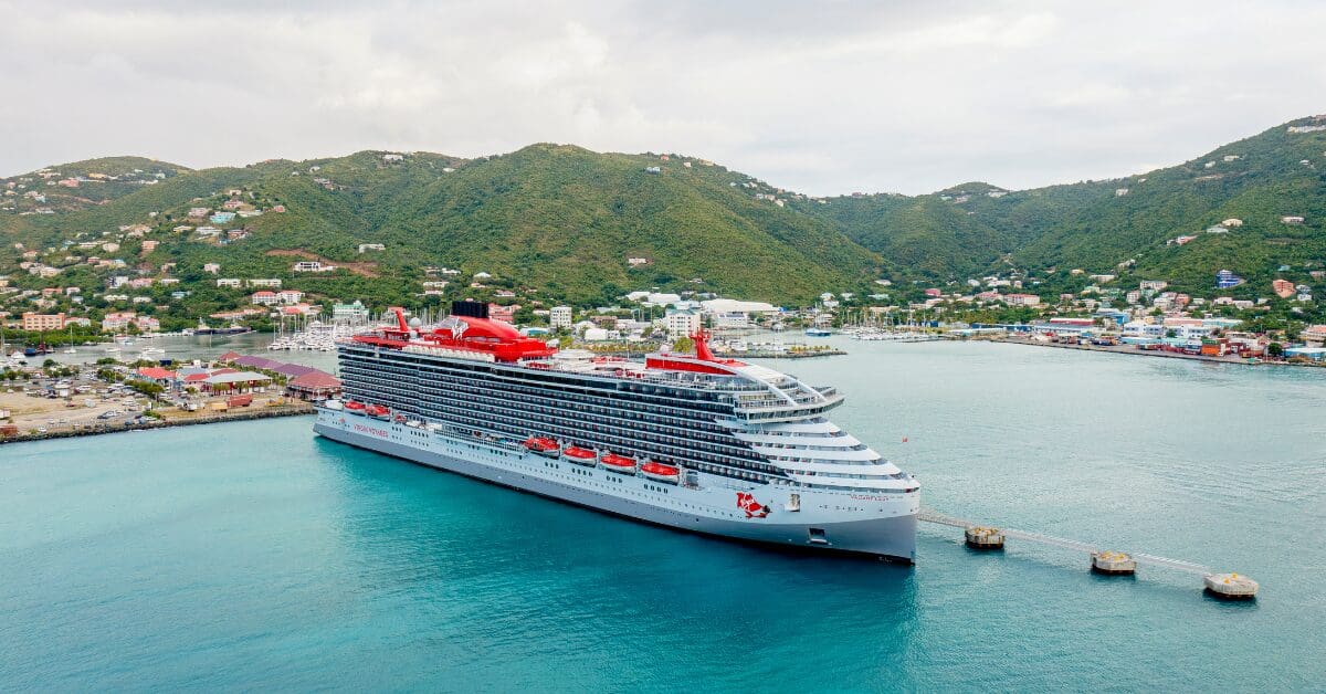 Virgin Voyages cruise ship docked at a Caribbean port with lush green hills and colorful buildings in the background.